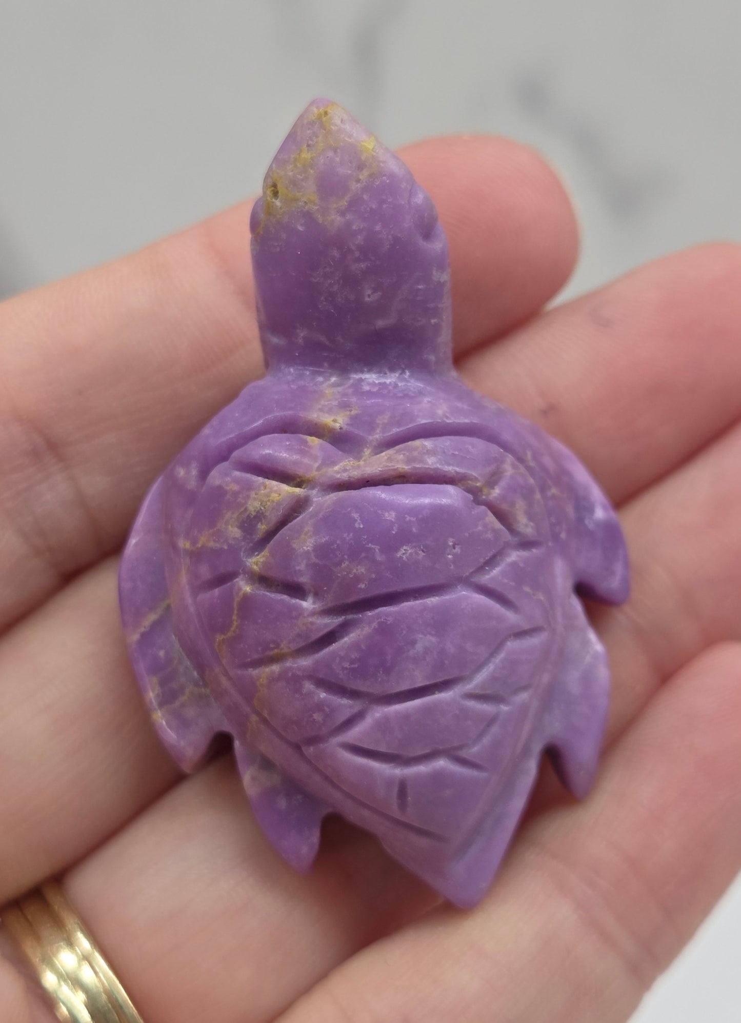 A carved purple turtle figurine resting on a person's hand, with a gold ring visible on the wrist against a light gray marble background.