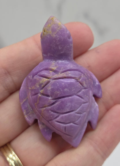 A carved purple turtle figurine resting on a person's hand, with a gold ring visible on the wrist against a light gray marble background.