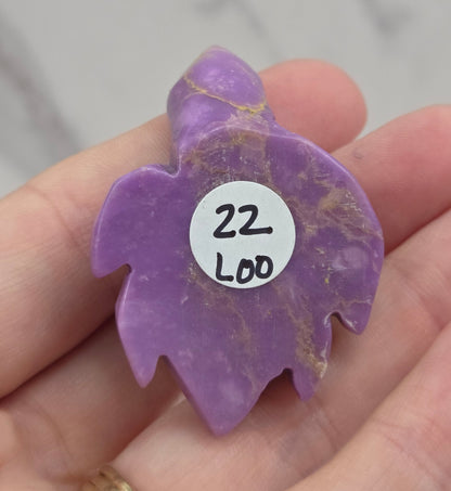A hand holds a small purple carved stone charm with gold veining and a white circular sticker that reads '22 Loo'.