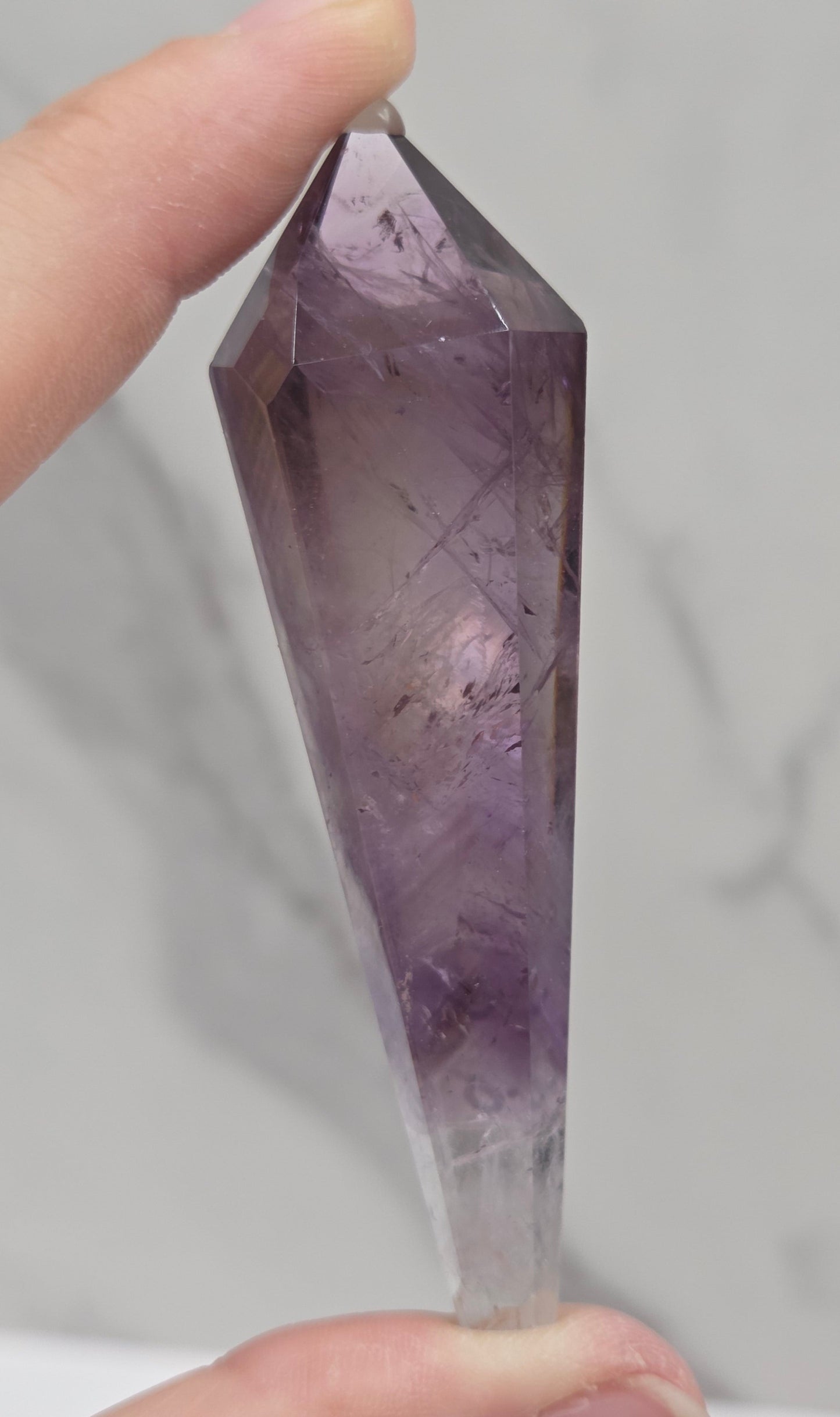 A hand holding a faceted purple crystal point against a light gray marble-patterned background.