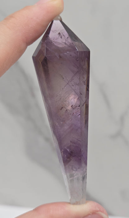 A hand holding a faceted purple crystal point against a light gray marble-patterned background.