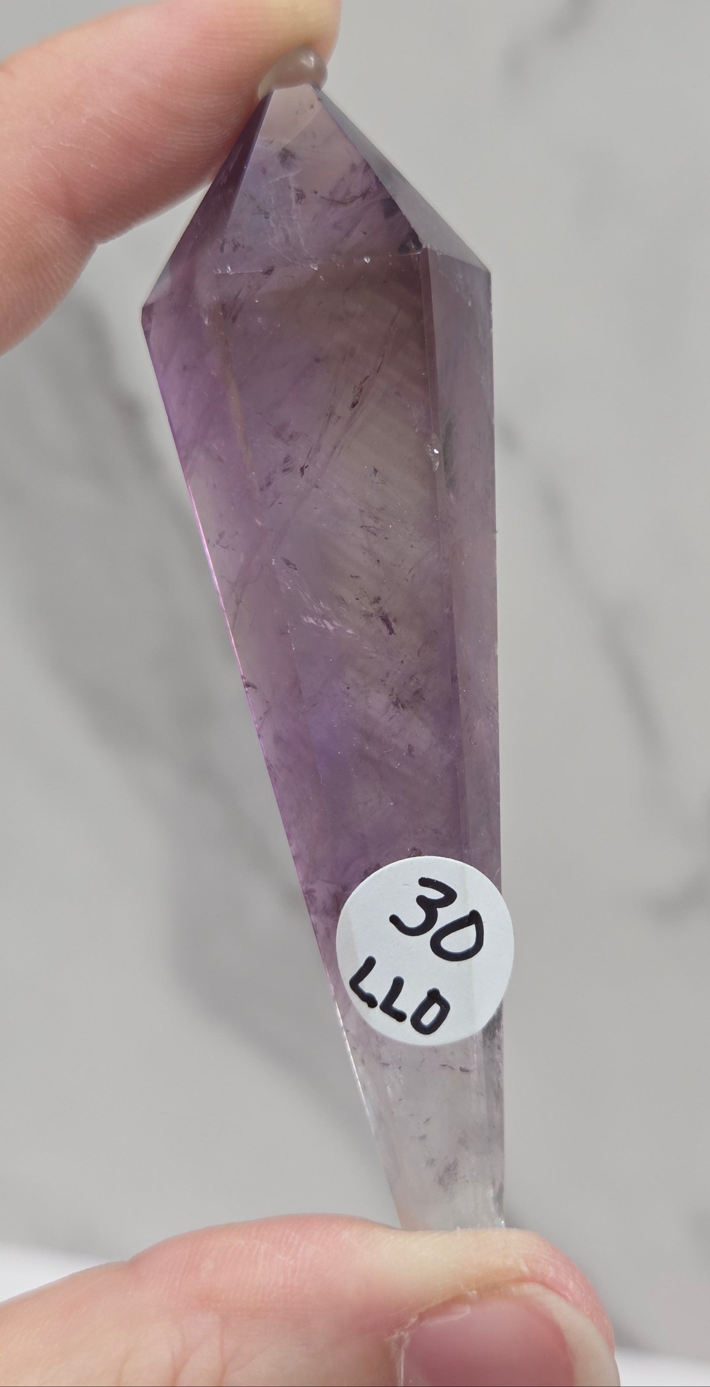 A hand holding a faceted purple crystal point, likely amethyst, with a small white sticker reading '3D LLD' on it, against a light marble-patterned background.