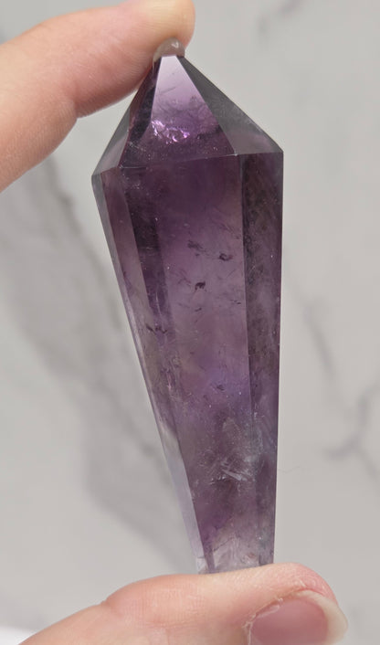 A hand holding a faceted purple crystal point against a light gray marble-patterned background.