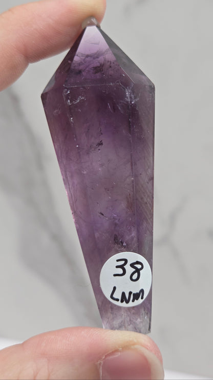 A hand holding a pointed amethyst crystal point against a light marble background. A small white sticker on the crystal reads '38 LMM'.