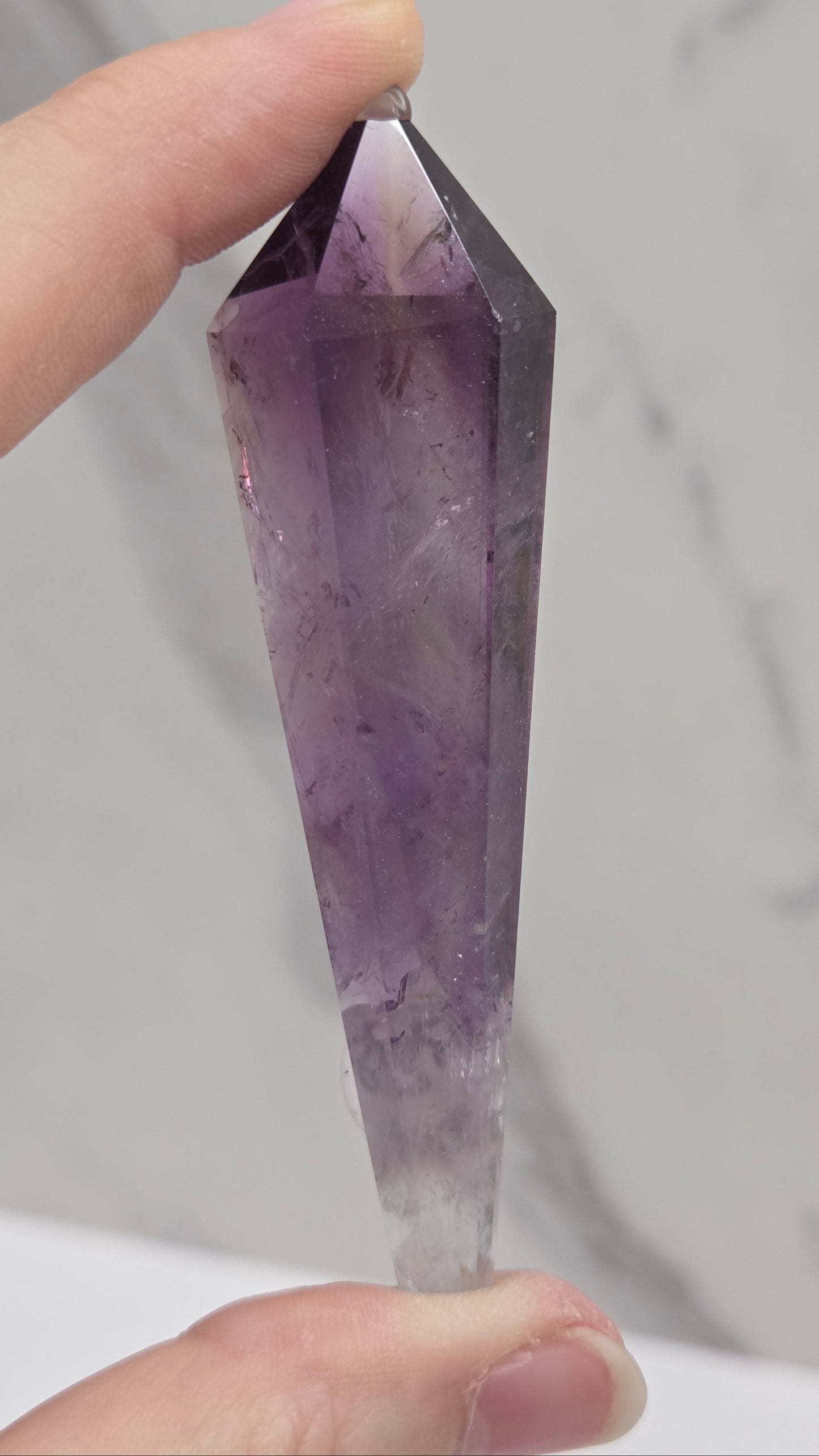 A faceted purple amethyst crystal point held between two hands against a light gray marble-patterned background.