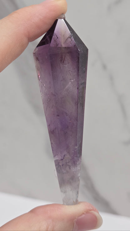 A faceted purple amethyst crystal point held between two hands against a light gray marble-patterned background.