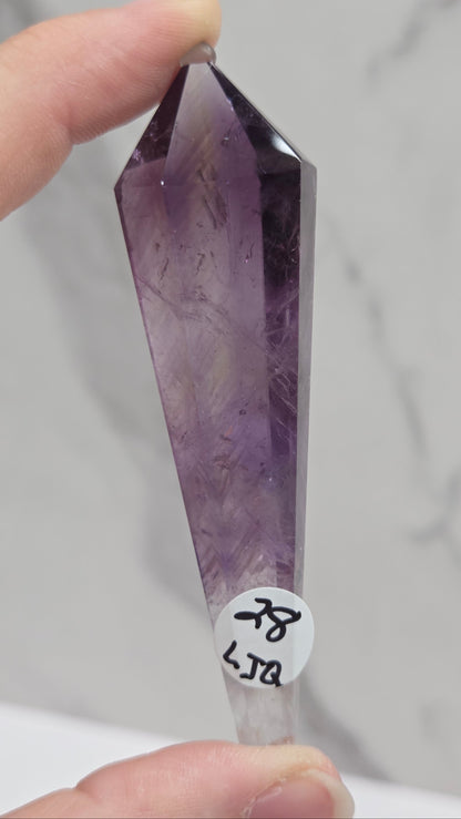 A hand holding a pointed amethyst crystal point with visible internal inclusions, against a light marble-like background. A small white label with black handwritten characters is attached near the base.