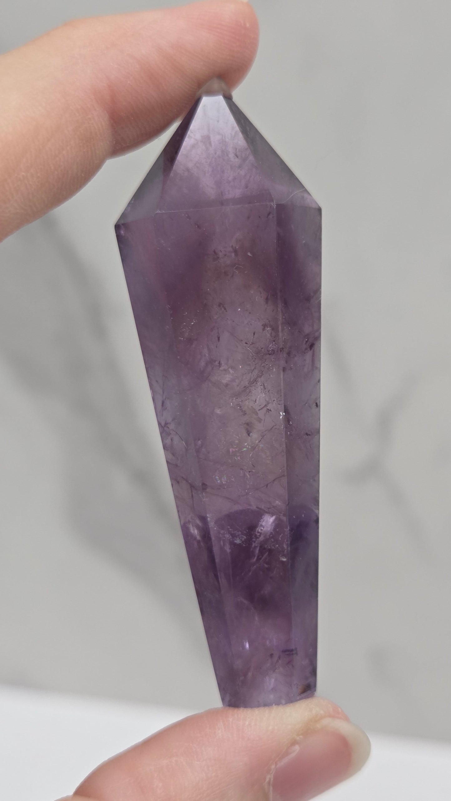 A faceted purple amethyst crystal point held between two hands against a light gray marble-patterned background.