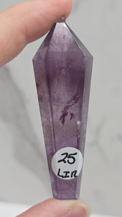 A hand holding a faceted purple amethyst crystal point against a light gray marble-patterned background. A small white circular sticker on the crystal reads '25 Lln'.