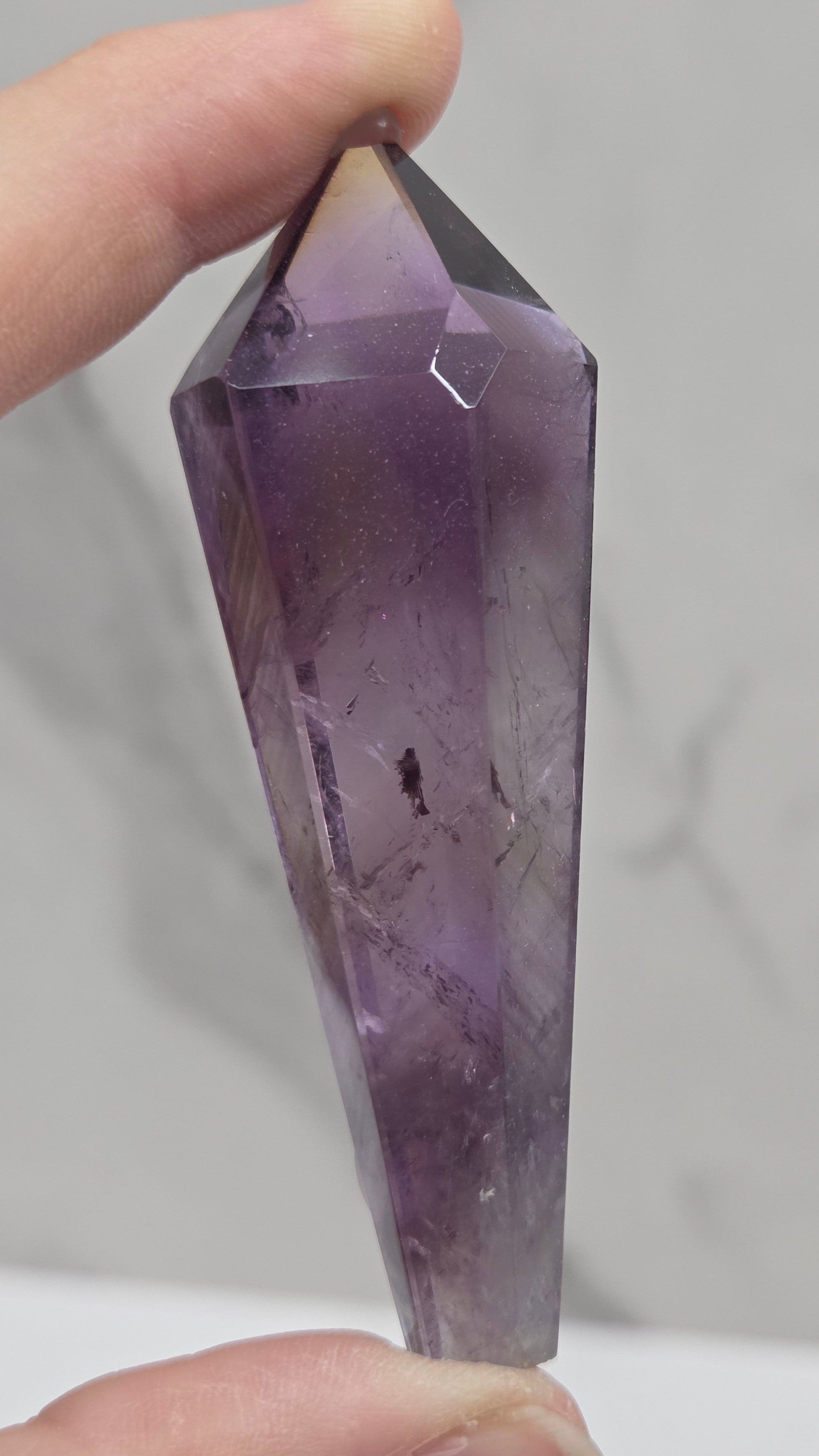 A faceted purple amethyst crystal point held between two hands against a light gray marble-patterned background.