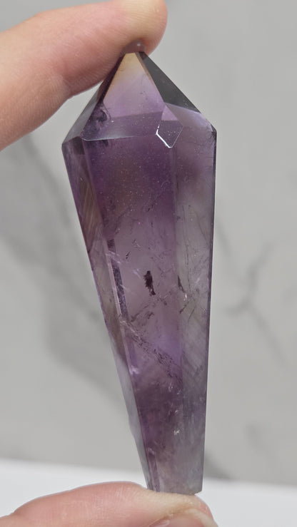 A faceted purple amethyst crystal point held between two hands against a light gray marble-patterned background.