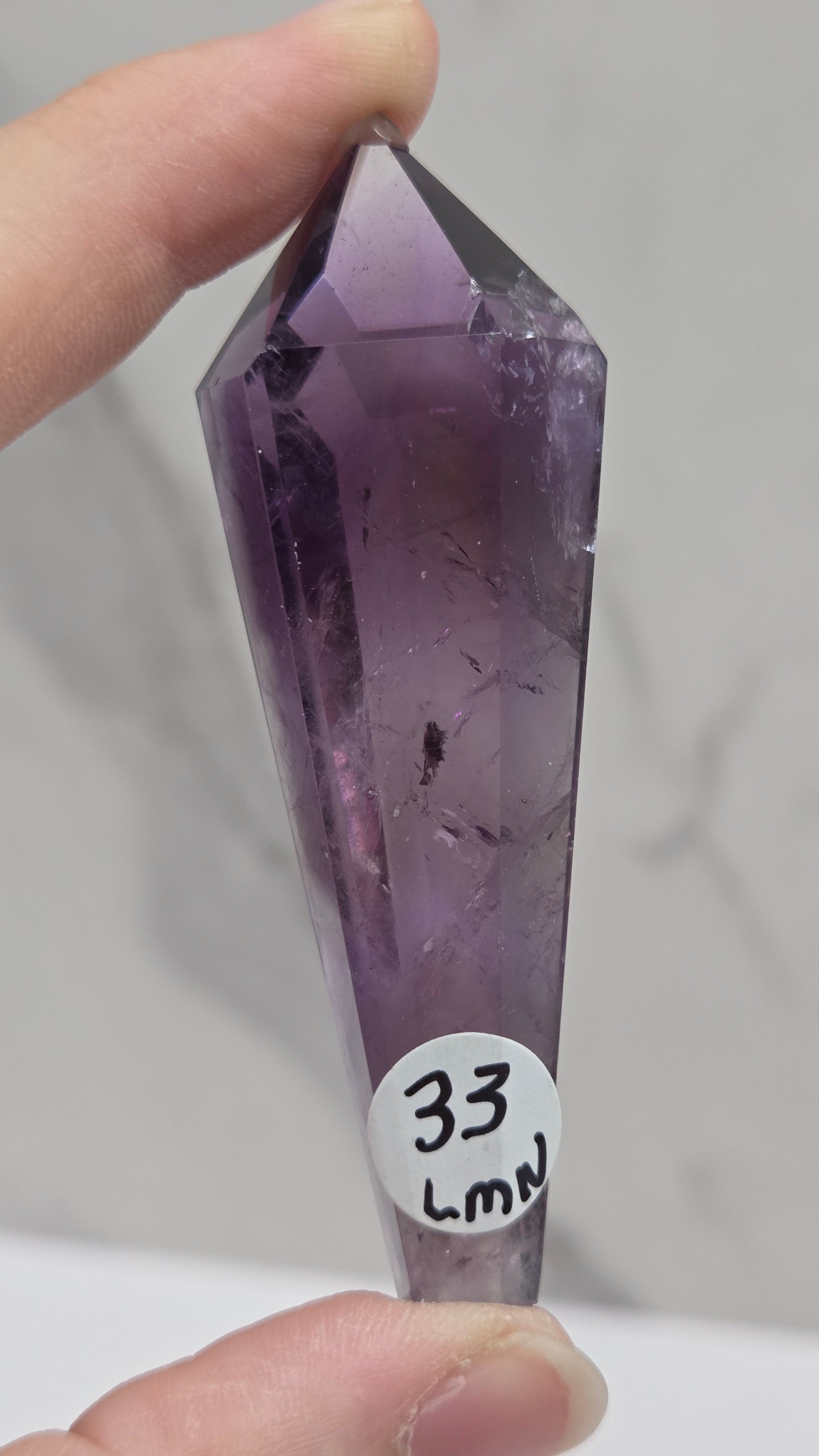 A hand holding a faceted purple amethyst crystal point against a light gray marble-patterned background. A small white circular sticker on the crystal reads '33 LMS'.