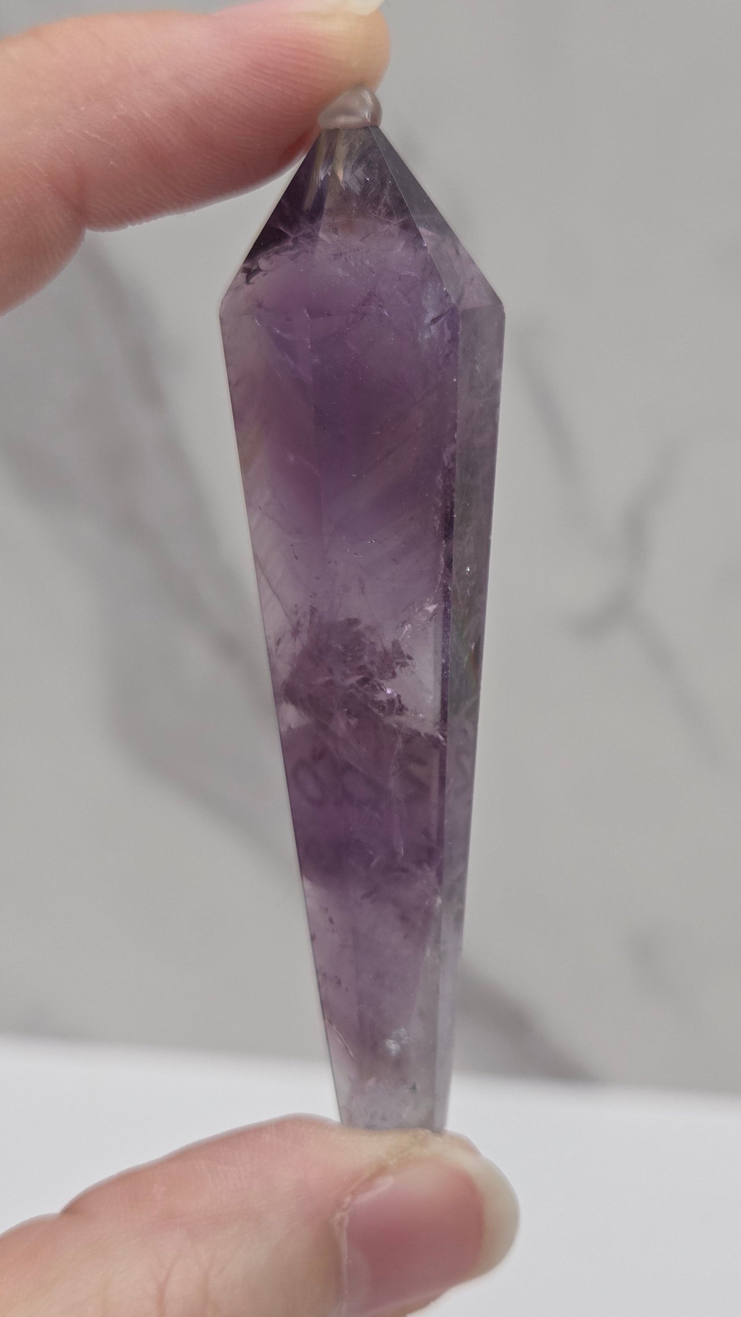 A hand holding a faceted purple amethyst crystal point against a light gray marble-patterned background.