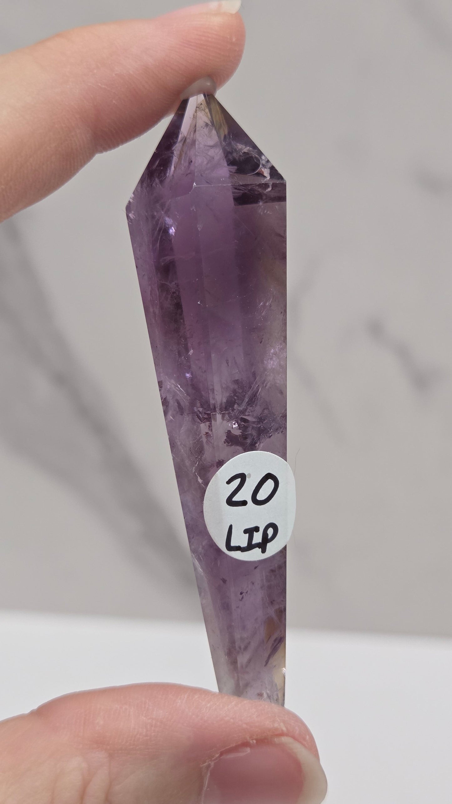 A hand holding a pointed amethyst crystal tower against a light gray marble-patterned background. A small white circular sticker with handwritten text is attached near the center of the crystal.