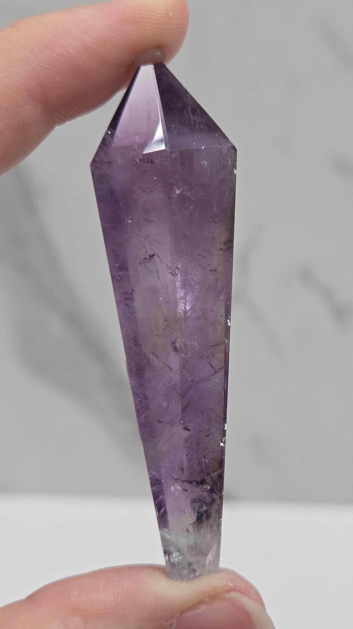 A faceted purple amethyst crystal point held between two fingers against a light gray marble-patterned background.