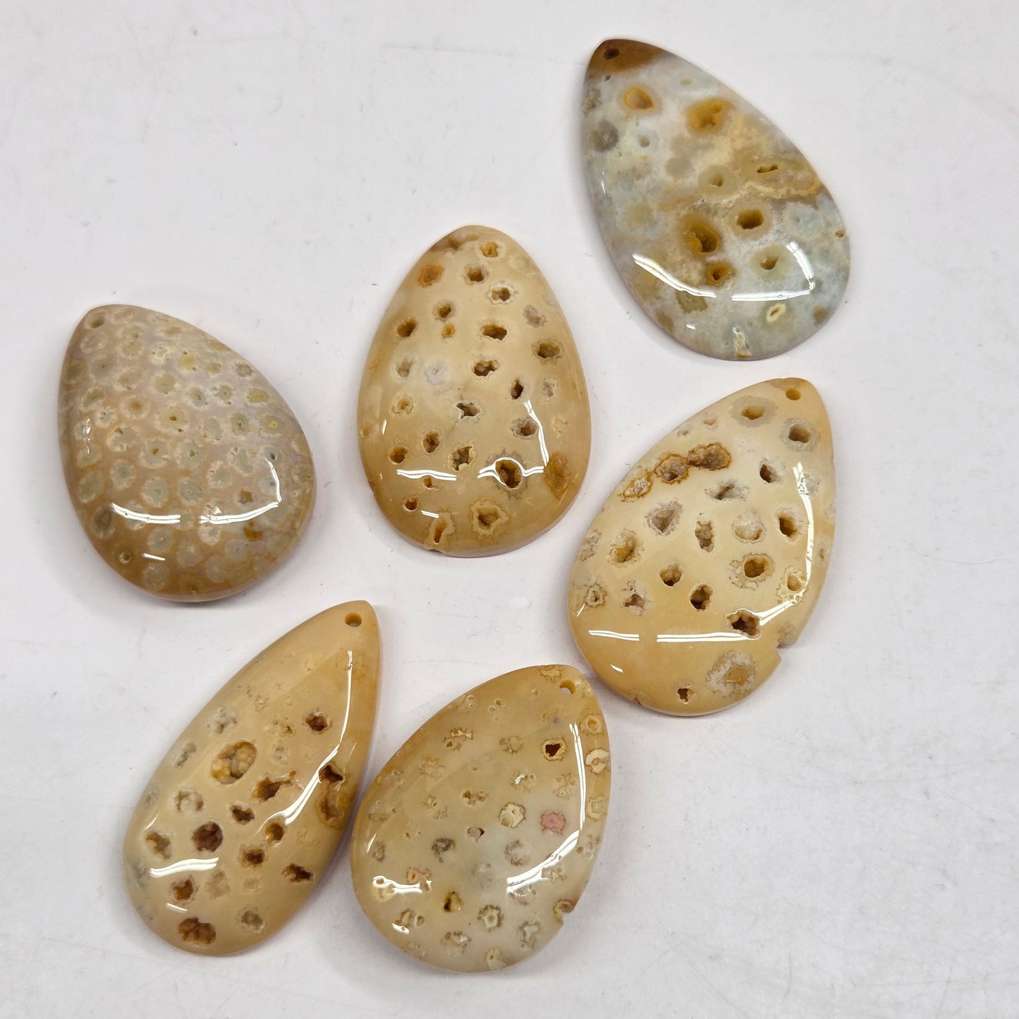 Six polished teardrop-shaped stone cabochons with a porous, sponge-like texture in shades of beige, tan, and light gray, arranged on a white background.