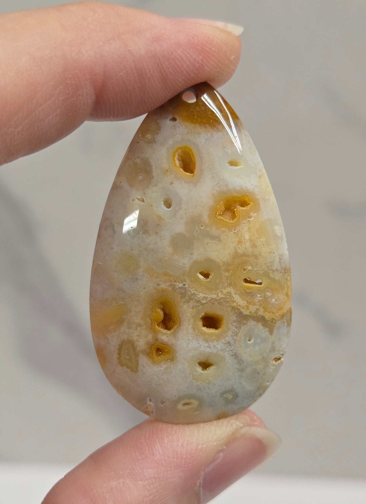 A hand holding a polished teardrop-shaped agate stone pendant with natural burl patterns in shades of beige, brown, and pale gray against a light gray marble background.