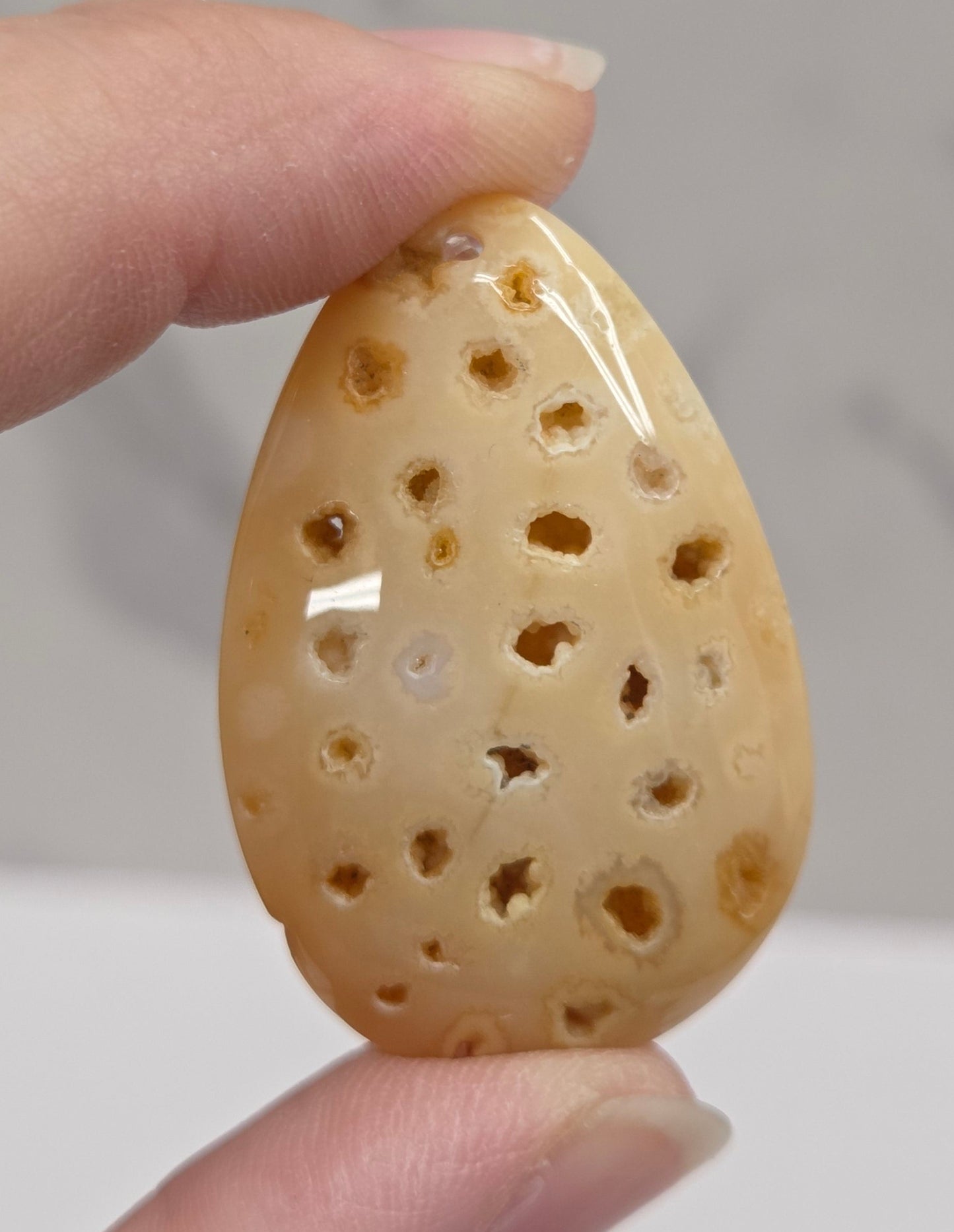 A hand holding a small, teardrop-shaped carved stone or bone ornament with multiple drilled holes, shown against a soft gray marble-patterned background.