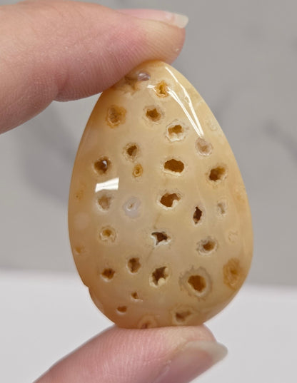 A hand holding a small, teardrop-shaped carved stone or bone ornament with multiple drilled holes, shown against a soft gray marble-patterned background.