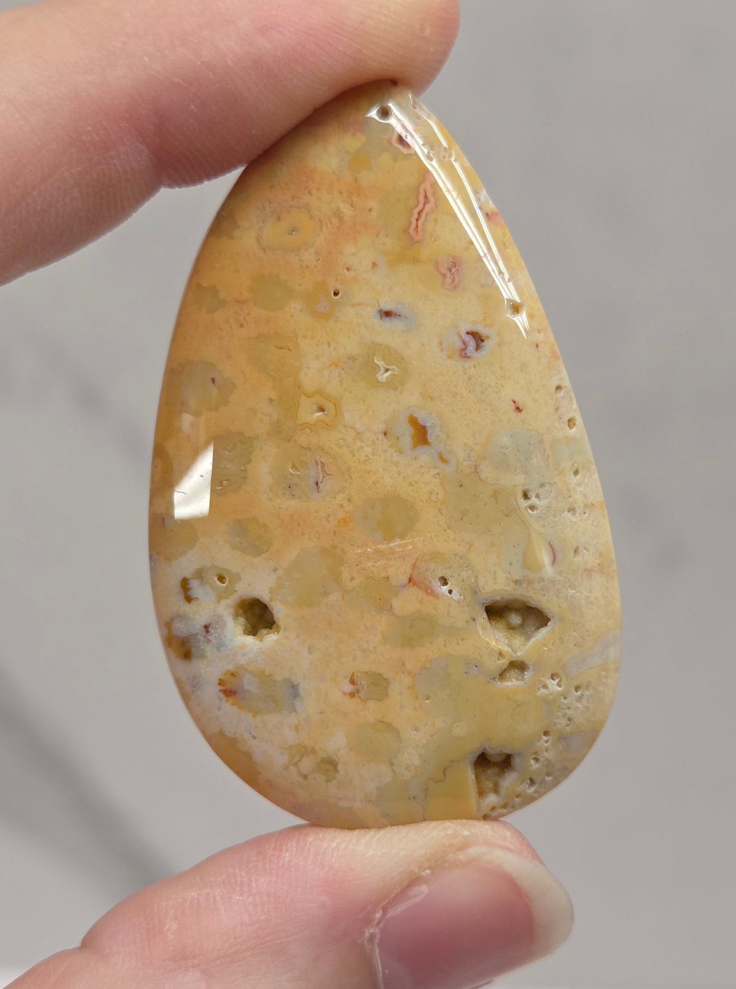 A hand holding a polished teardrop-shaped stone cabochon with a honeycomb-like porous texture in shades of beige, tan, and light brown, set against a soft gray marble background.