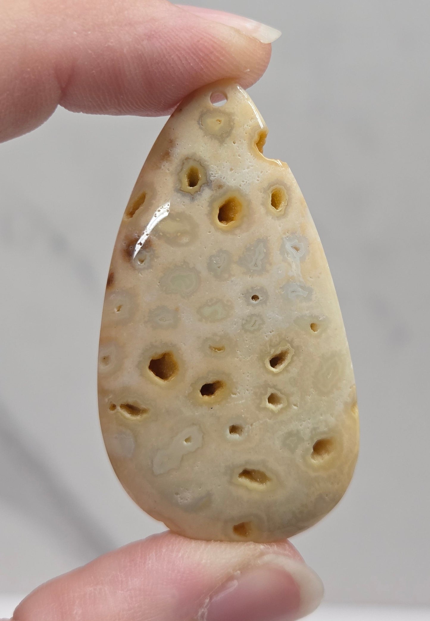 A hand holding a teardrop-shaped carved stone pendant with a honeycomb-like pattern of holes, shown against a light gray marble background.