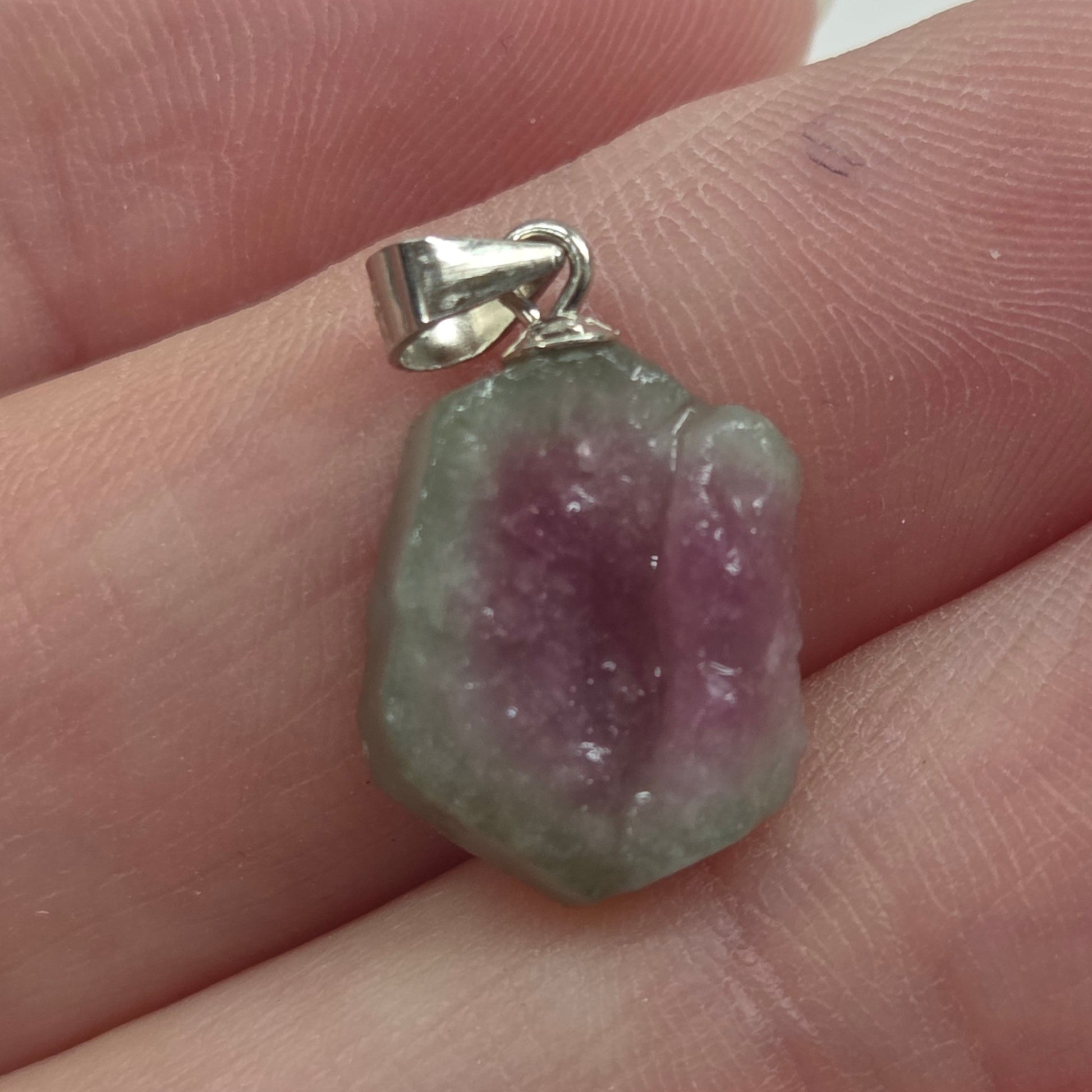 A small raw gemstone pendant with a silver bail, resting on a person's fingers. The stone is characterized by a distinct color gradient transitioning from a dusty purple center to a light green outer edge, set in a simple silver clasp.