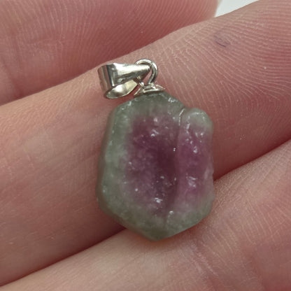A small raw gemstone pendant with a silver bail, resting on a person's fingers. The stone is characterized by a distinct color gradient transitioning from a dusty purple center to a light green outer edge, set in a simple silver clasp.