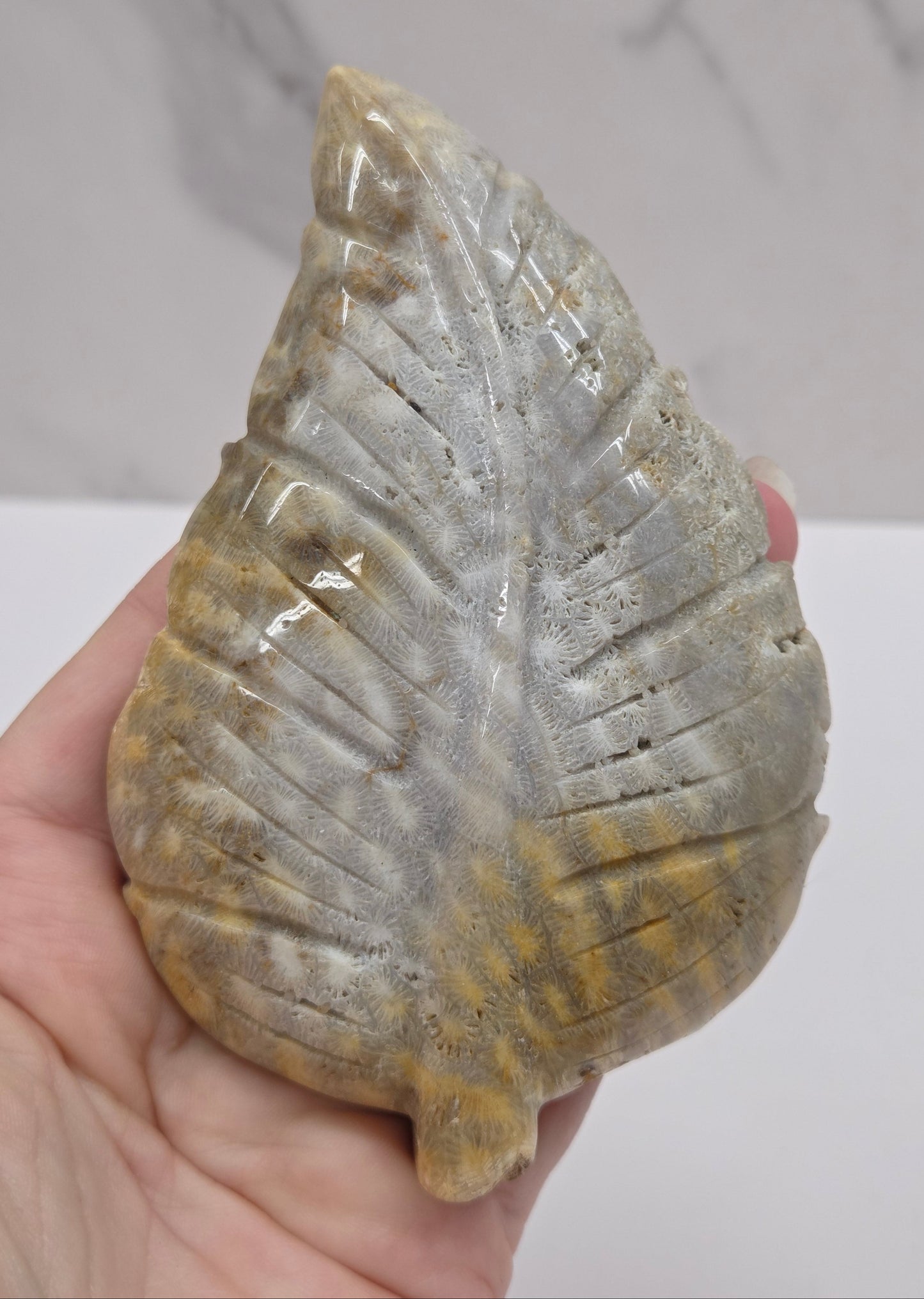 A hand holding a polished carved stone leaf ornament with a textured, ribbed surface in gray, beige, and golden-brown tones, set against a light gray marble-patterned background.