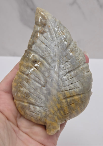 A hand holding a polished carved stone leaf ornament with a textured, ribbed surface in gray, beige, and golden-brown tones, set against a light gray marble-patterned background.