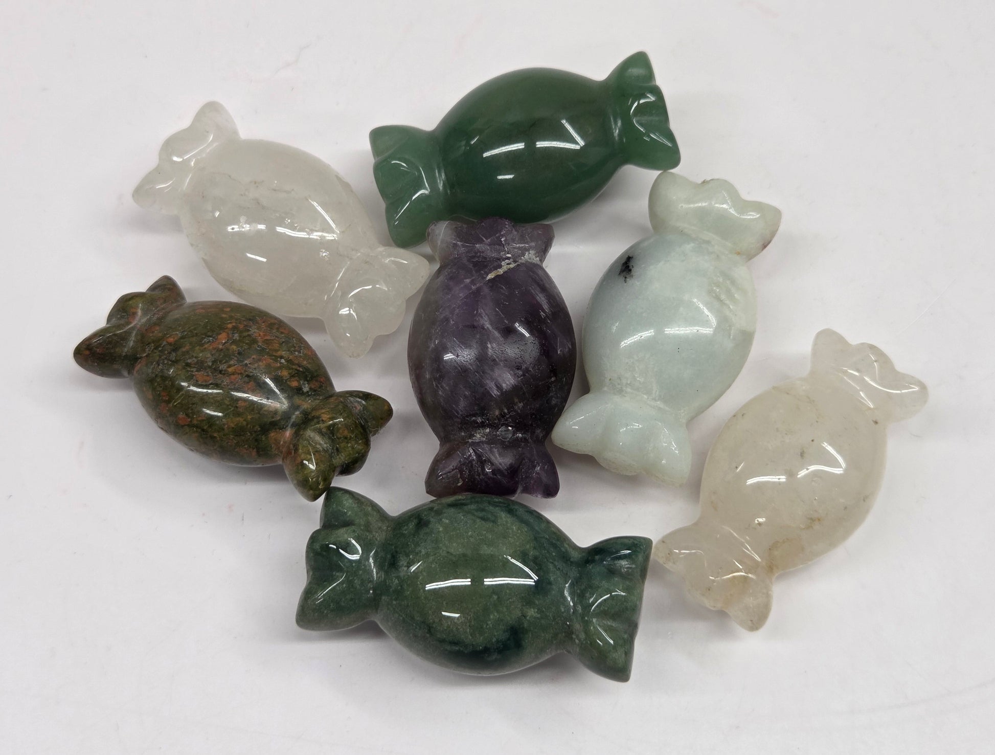 A set of seven polished stone candy-shaped figurines in various colors including green, purple, white, and brown, arranged on a plain white surface.