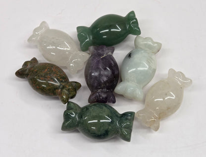 A set of seven polished stone candy-shaped figurines in various colors including green, purple, white, and brown, arranged on a plain white surface.