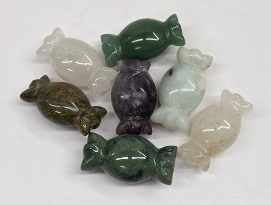 A set of seven polished stone candy-shaped figurines in various colors including green, purple, white, and brown, arranged on a plain white surface.