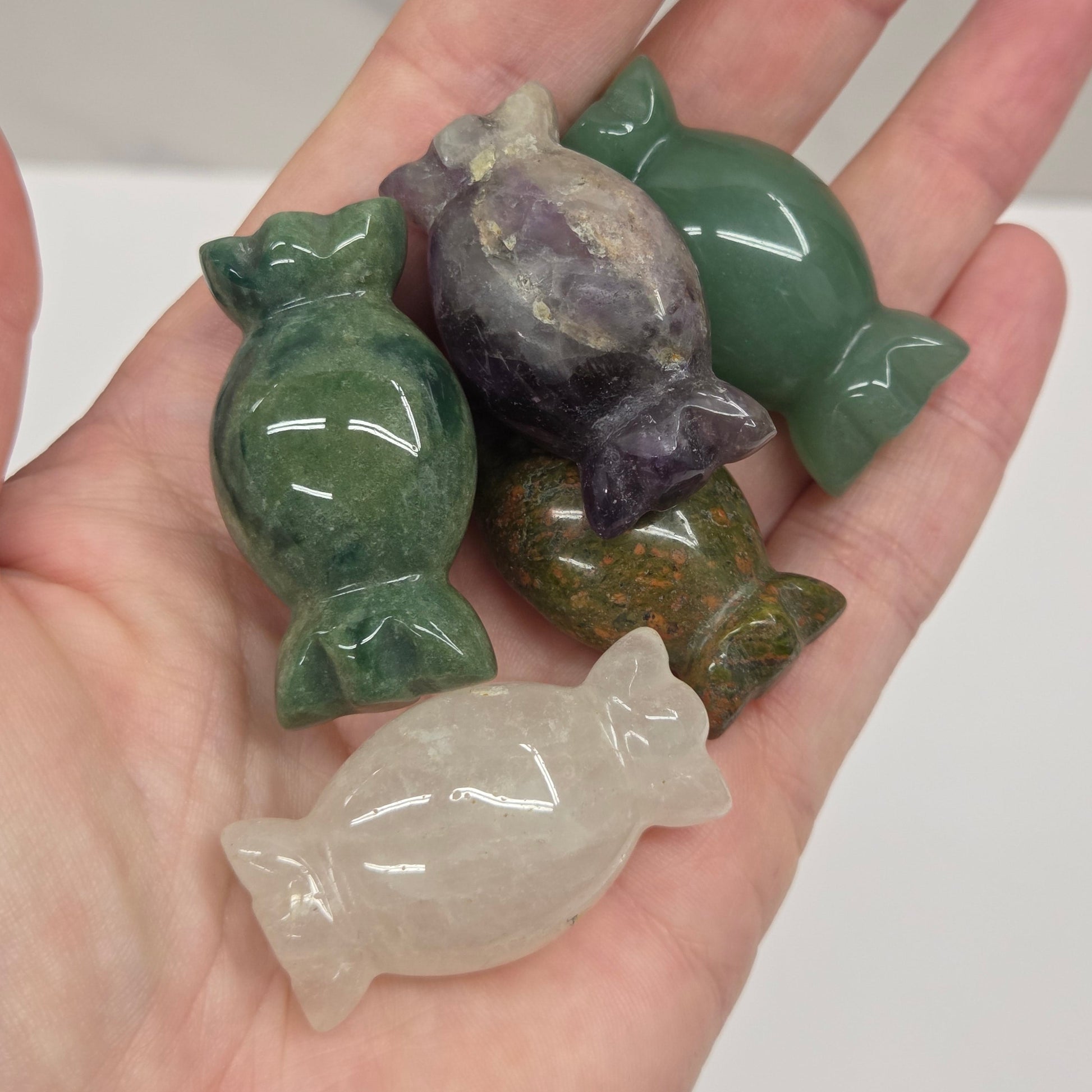Five small polished stone figurines shaped like wrapped candies are displayed on an open hand. The set includes green, purple, olive green, and milky white stones with a glossy finish.