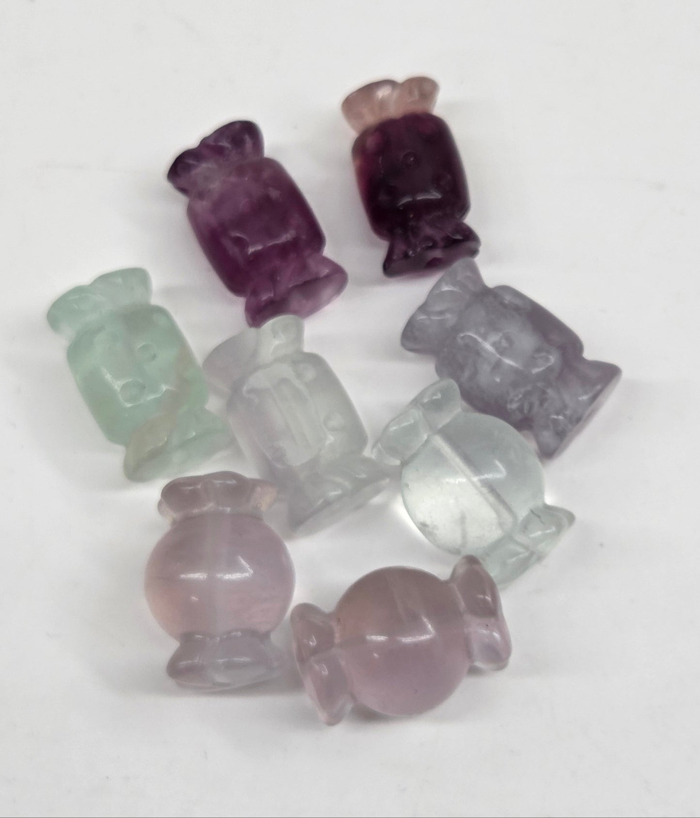 A group of small carved gemstone beads shaped like candies, shown on a plain white background. The beads are translucent and come in shades of purple, pale pink, light green, and clear.