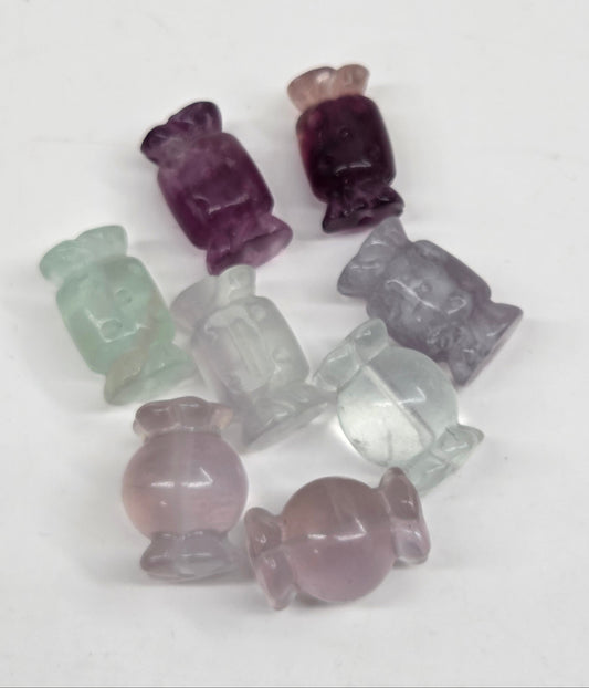 A group of small carved gemstone beads shaped like candies, shown on a plain white background. The beads are translucent and come in shades of purple, pale pink, light green, and clear.
