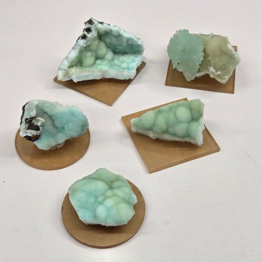 Five pieces of pale mint-green crystal or mineral specimen with rough, bubbly textures, each mounted on a small brown cardboard backing, arranged on a plain white surface.