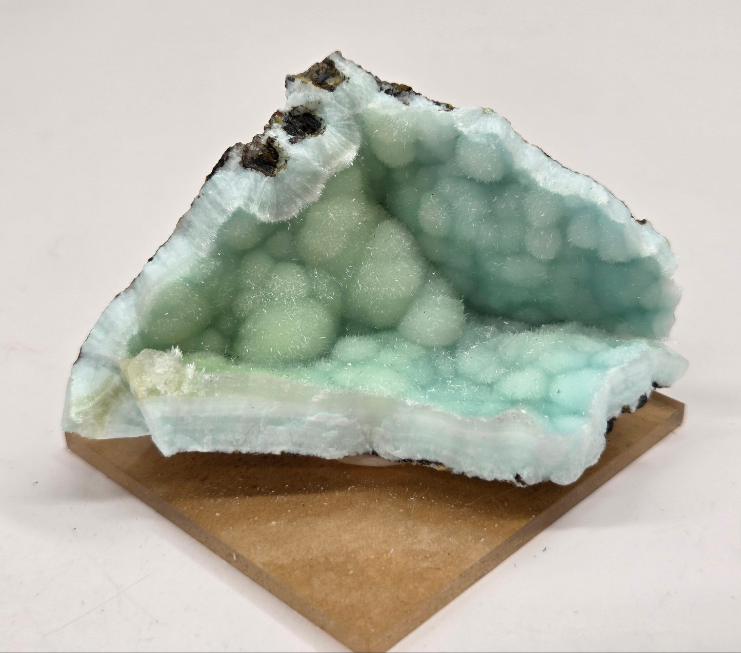 A light blue-green crystal geode specimen with bubbly druzy formations, mounted on a square tan cardboard display block against a plain off-white background.