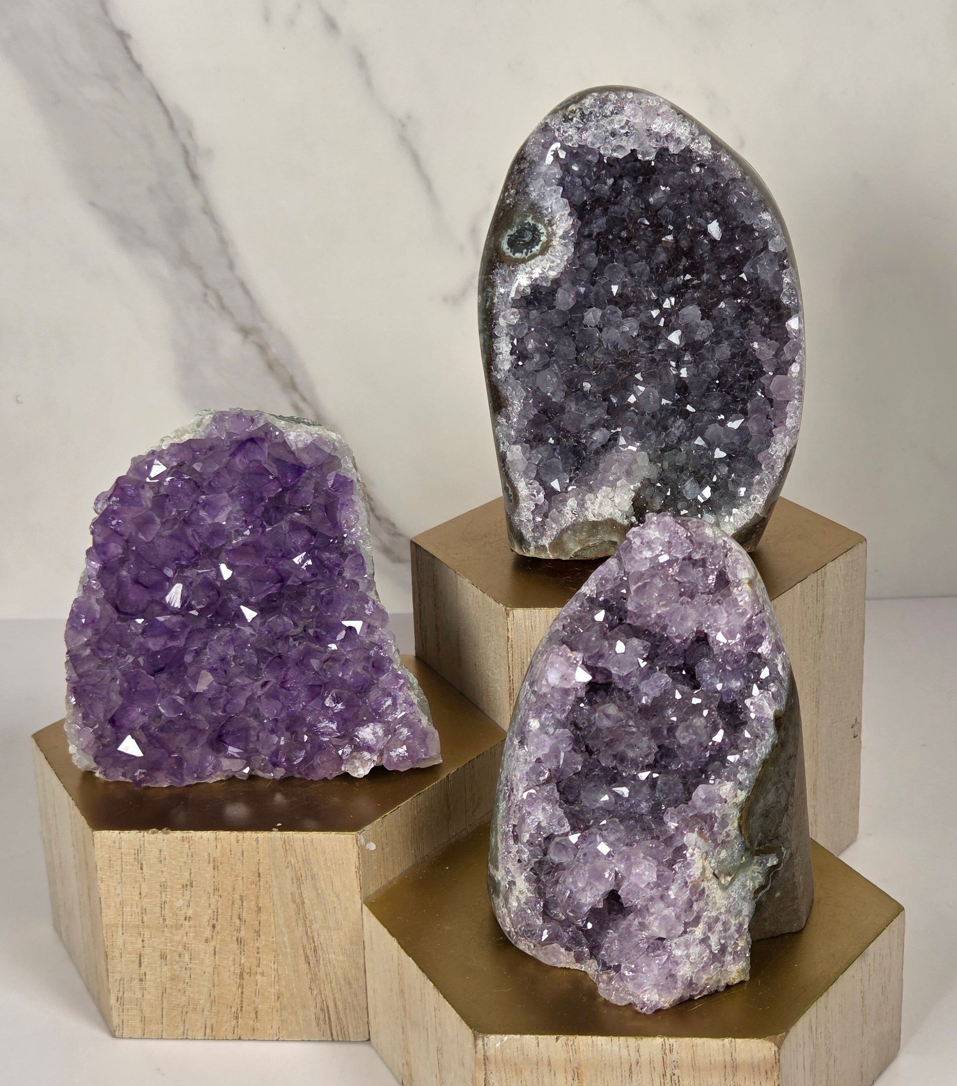 Three raw amethyst geode clusters in different sizes mounted on a light wood cross-shaped display stand against a white marble-patterned wall.