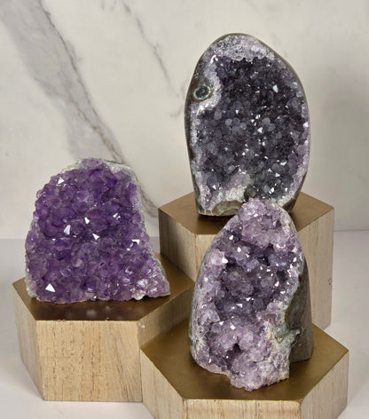 Three raw amethyst geode clusters in different sizes mounted on a light wood cross-shaped display stand against a white marble-patterned wall.
