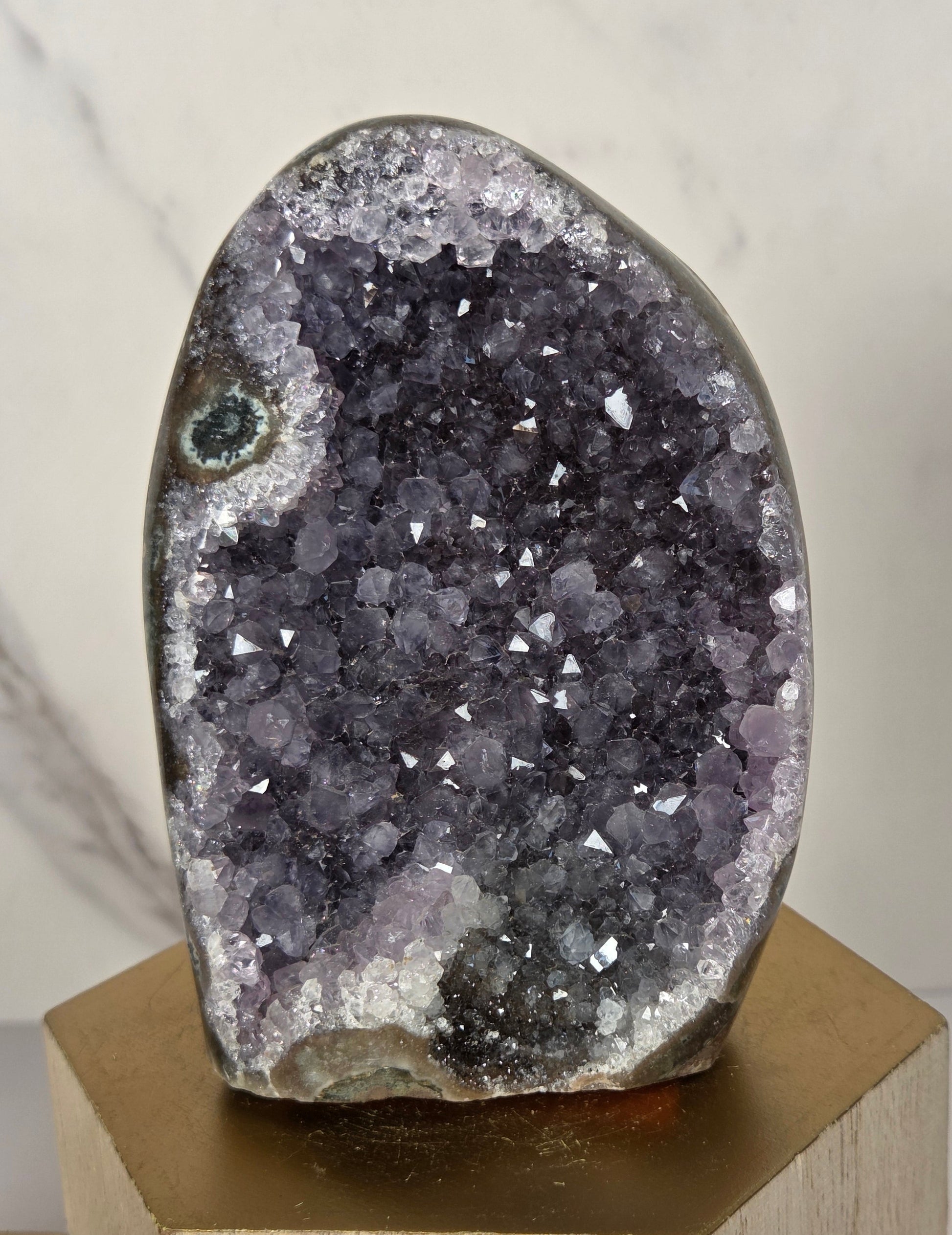 A polished geode specimen with a dark purple and gray crystal cavity, mounted on a light wood block against a white marble background.