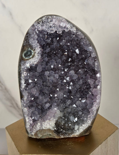 A polished geode specimen with a dark purple and gray crystal cavity, mounted on a light wood block against a white marble background.