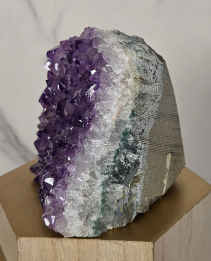 A natural amethyst crystal cluster geode mount displayed on a light wood geometric block against a white marble background.