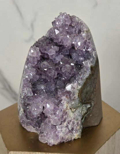 A raw amethyst crystal cluster with prominent purple and lavender crystalline formations, mounted on a light brown wooden display block against a white marble-patterned background.