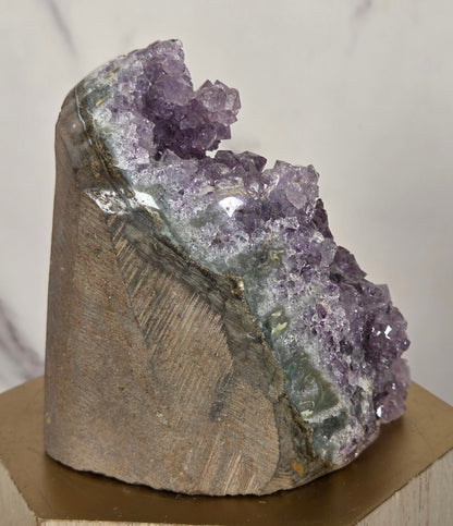 A raw amethyst crystal cluster mounted on a hexagonal wooden display block, photographed against a light marble-patterned background.