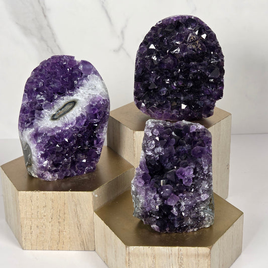 Three purple amethyst geode clusters mounted on a light beige wooden geometric display stand against a white marble background.