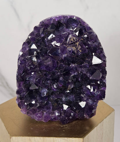 A purple amethyst crystal cluster geode mounted on a light wood block, set against a white marble background.