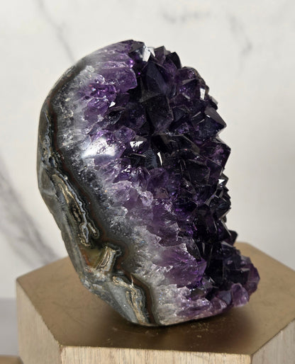An amethyst crystal geode cluster mounted on a light wood block, photographed against a white marble-patterned background.