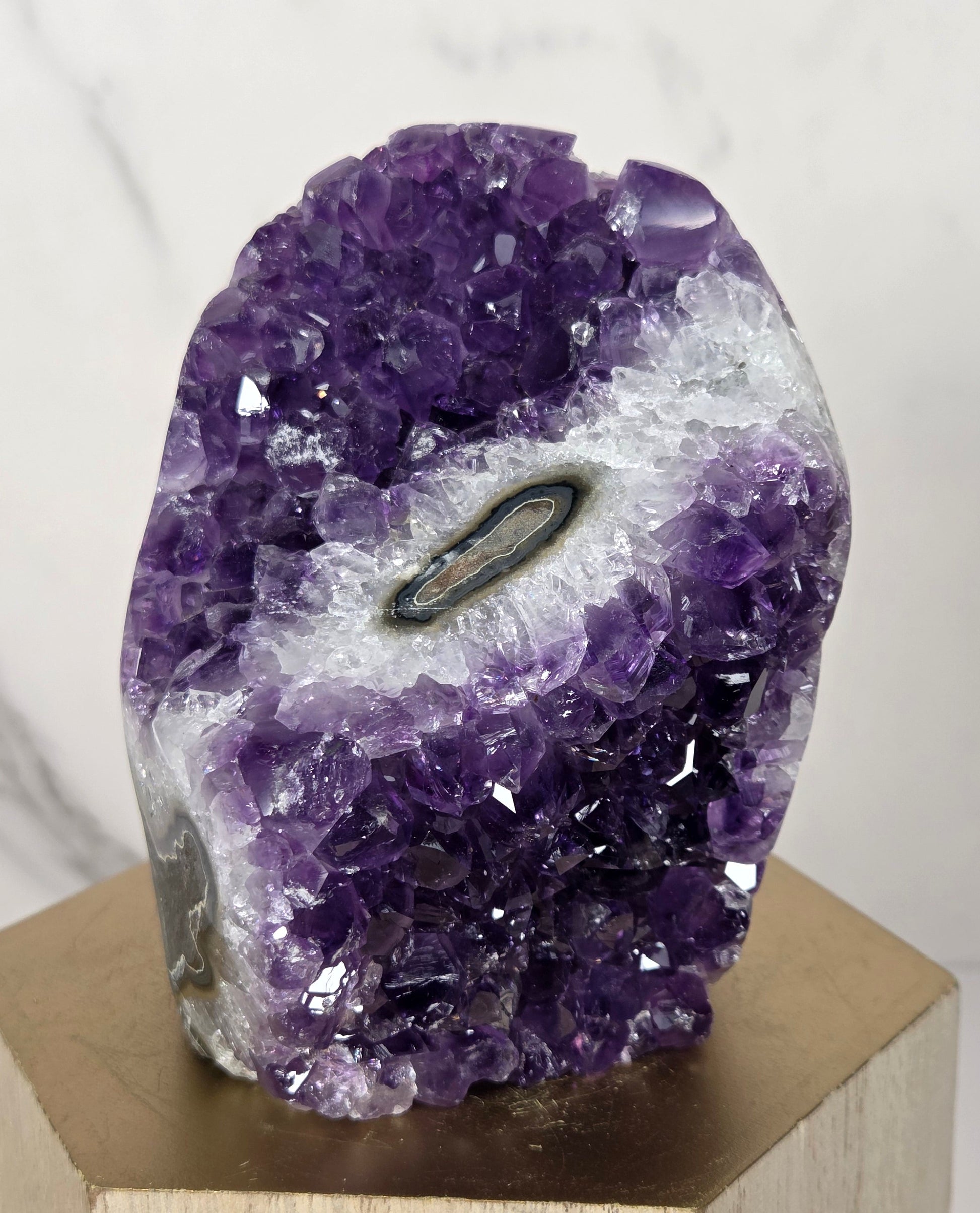 A natural amethyst geode specimen with a central black tourmaline tube, mounted on a light-colored wooden display block against a white marble background.