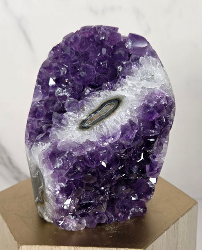 A natural amethyst geode specimen with a central black tourmaline tube, mounted on a light-colored wooden display block against a white marble background.