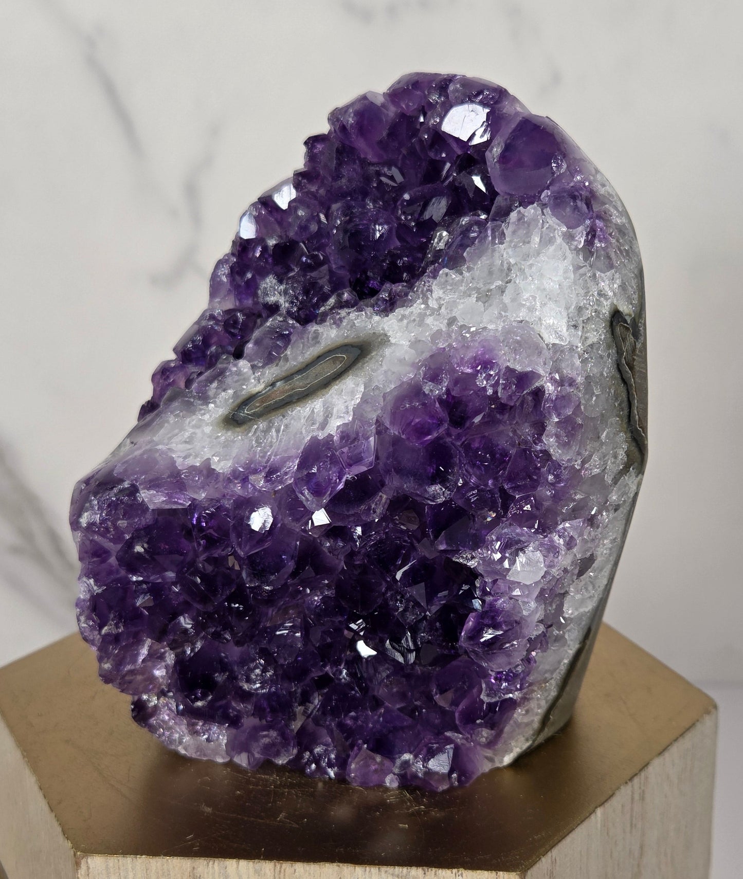 A raw amethyst geode cluster with a white matrix and dark matrix lines, mounted on a hexagonal wooden display block against a white marble background.
